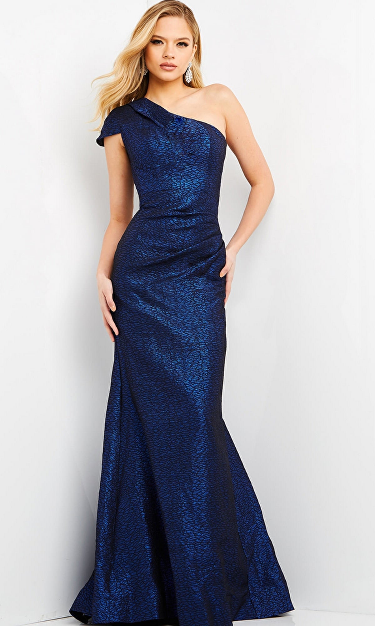 A woman wearing the Jovani 06751 One-Shoulder Metallic Formal Dress in blue, featuring a subtle textured pattern, stands against a plain light background.
